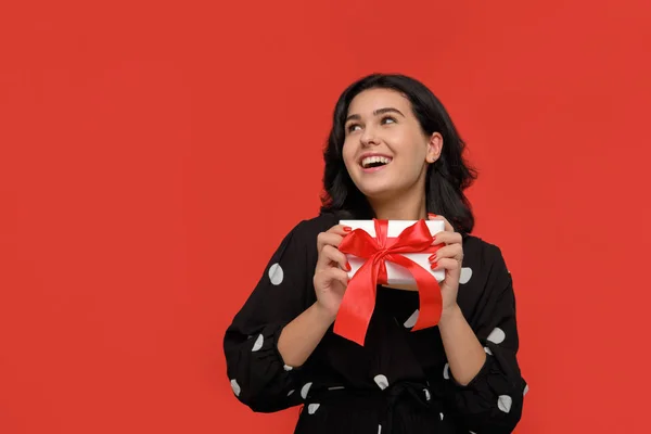 Brunetka kobieta w czarnej sukience uśmiechnięta gospodarstwa Christmas Giftbox w kolorze białym z czerwoną wstążką. — Zdjęcie stockowe