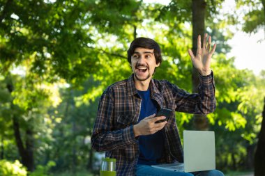 Sakallı ve bıyıklı genç bir serbest yazar. Parkta dizüstü bilgisayarla oturuyor ve elinde akıllı telefon var..