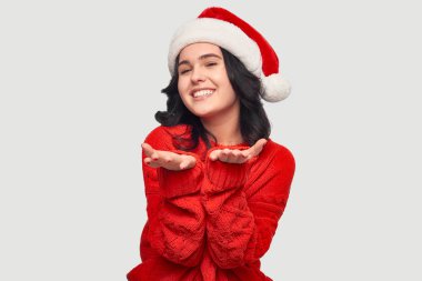Kırmızı kazaklı ve Noel Baba şapkalı kız gri arka planda izole bir şekilde kameraya hava öpücüğü gönderiyor.