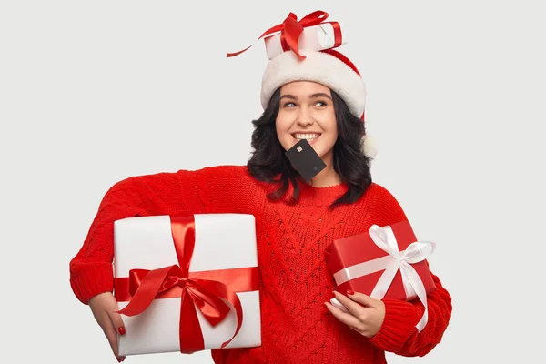 身穿红色毛衣、头戴圣诞礼帽、头戴银行卡的女孩，手里拿着圣诞礼品盒，牙齿塞满银行卡 — 图库照片