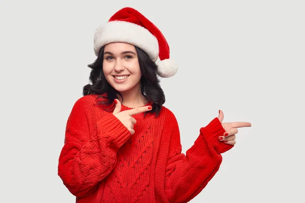 Hübsches Mädchen in rotem Pullover und Weihnachtsmütze, das auf den Kopierraum vor grauem Hintergrund zeigt — Stockfoto