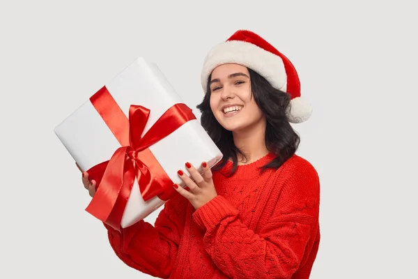 Chica morena en un suéter rojo y sombrero de Santa Claus sosteniendo caja de regalo blanca con cinta roja —  Fotos de Stock