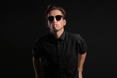 Siyah tişörtlü ve güneş gözlüklü şık genç adam siyah arka planda sigara içiyor..