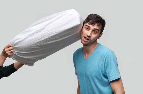 Pretty brunet man in a blue tee is shocked because white pillow flying in his dirrection. — Stock Photo, Image