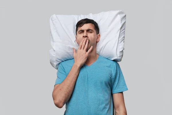 Hombre moreno cansado en una camiseta azul descansando sobre una almohada blanca aislada sobre fondo gris . — Foto de Stock