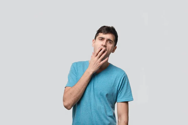 Young man with white feather from a pillow yawning over grey background. Tired after long day — Stock Photo, Image
