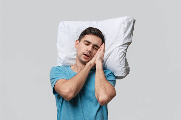 Tired brunet man in a blue tee resting on a white pillow isolated over grey background.