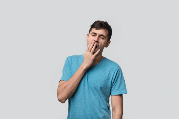 Genç esmer adam gri arka planda tek başına esniyor. Uzun bir çalışma gününden sonra yorgun olmak.