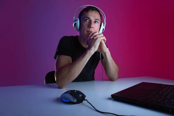 Задумчивый геймер в наушниках делает перерыв во время видеоигры в темной комнате, освещенной неоновыми огнями . — стоковое фото