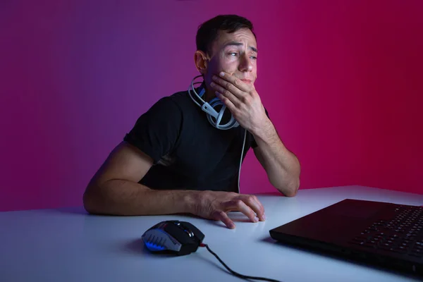 Скептик-геймер в наушниках играет в видеоигры на компьютере в темной комнате, освещенной неоновыми огнями — стоковое фото