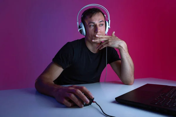 Escéptico jugador con auriculares jugando videojuegos en el ordenador en una habitación oscura iluminada con luces de neón . —  Fotos de Stock