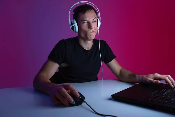 Escéptico jugador con auriculares jugando videojuegos en el ordenador en una habitación oscura iluminada con luces de neón —  Fotos de Stock