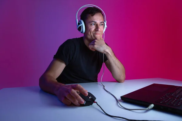 Sceptyczny gracz noszący słuchawki grający w gry wideo na komputerze w ciemnym pokoju oświetlonym światłami neonowymi. — Zdjęcie stockowe