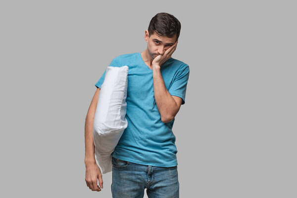 Tired brunet man in a blue tee resting on a white pillow isolated over grey background.