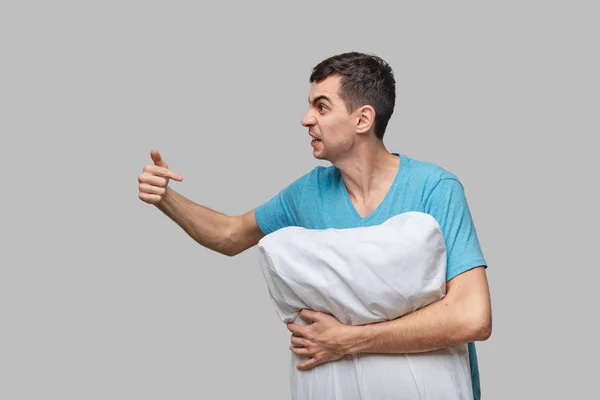 Hombre moreno cansado en una camiseta azul apuntando a la almohada blanca aislada sobre fondo gris . — Foto de Stock