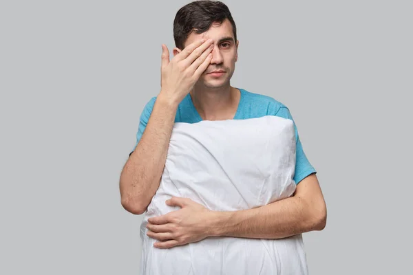 Young brunet man rubbing eyes holding white pillow