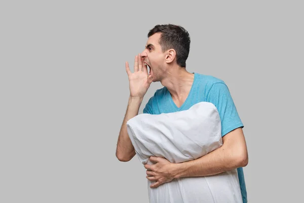 Hombre morena cansado en una camiseta azul grito sosteniendo almohada blanca aislada sobre fondo gris . — Foto de Stock