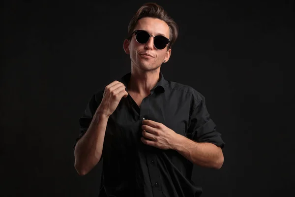 Confident stylish man in black eyeglasses buttoning up balck shirt isolated over black background.