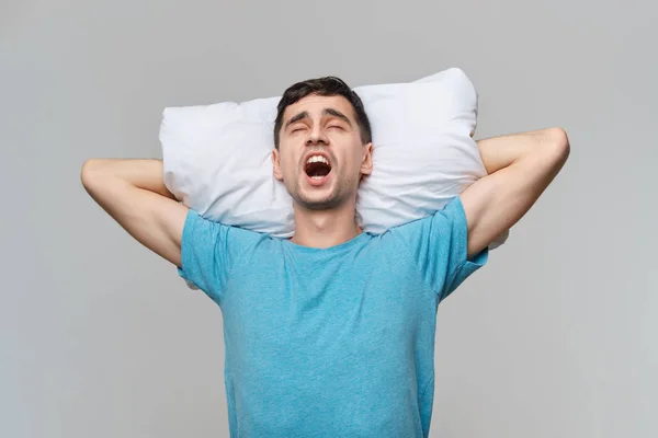 Hombre moreno cansado en una camiseta azul descansando sobre una almohada blanca aislada sobre fondo gris . — Foto de Stock