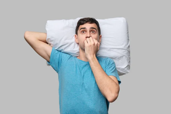 Hombre morena cansado en una camiseta azul está asustado acostado sobre una almohada blanca aislado sobre fondo gris . — Foto de Stock