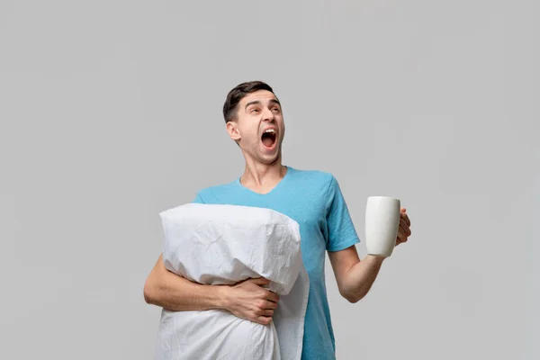 Junger brünetter Mann gähnt mit weißer Tasse Kaffee in der einen und weißem Kissen in der anderen Hand. — Stockfoto