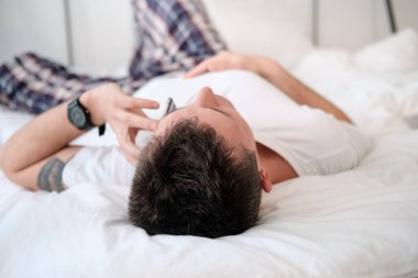 Beyaz gömlekli ve pijamalı yakışıklı bir genç yatakta uzanıyor ve telefonda gülümsüyor.