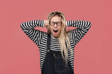 Çizgili gömlekli ve tulumlu genç bir kız iki eliyle başını tutarak bağırıyor.