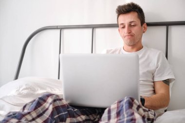 Beyaz çarşaflı beyaz gömlekli genç adam dizüstü bilgisayara bakarken üzgün..