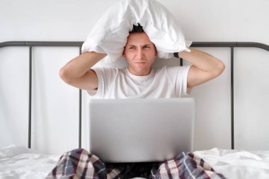 Angry young man in the white shirt and pajama holding white pillow on a head and laptop on knees