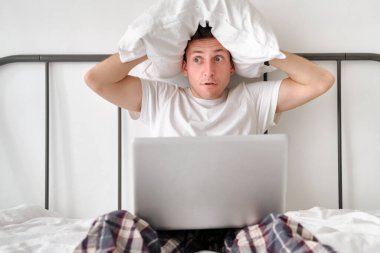 Angry young man in the white shirt and pajama holding white pillow on a head and laptop on knees