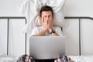 Angry young man in the white shirt and pajama holding white pillow on a head and laptop on knees