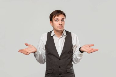 Young man in a white shirt and grey vest shrugging his shoulders expressing emotion of confusion.