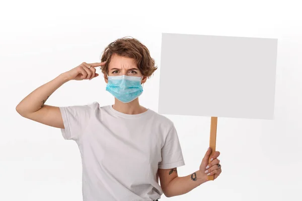 Worried woman wearing blue medical mask holds picket sign with copy space and piniting at her head — ストック写真