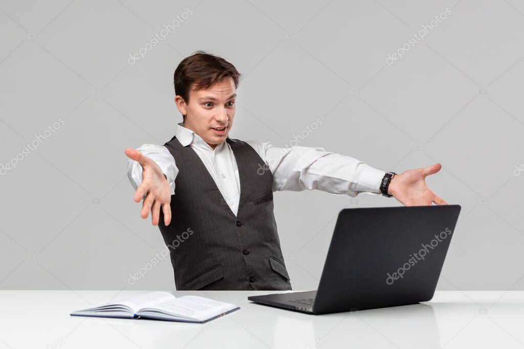 Man in a white t-shirt and grey vest is shocked looking at the laptop on a white table