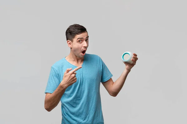 Young brunet man is shocked holding alarm-clock isolated over grey background — ストック写真