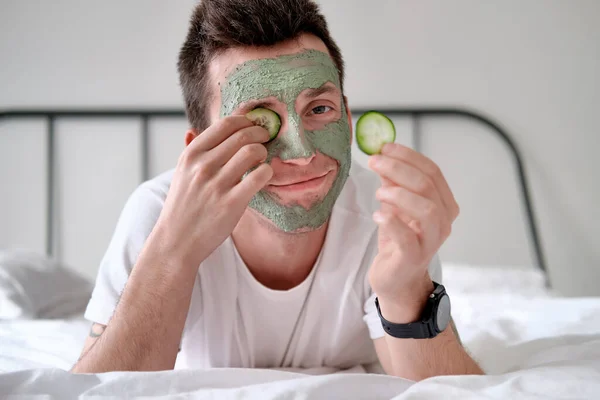 Young man in a white shirt with applied green cosmetic mask holding pieces of cucumber