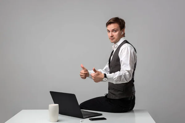 Бізнесмен у білій футболці та сірому жилеті тримає великі пальці вгору, сидячи на білому столі — стокове фото