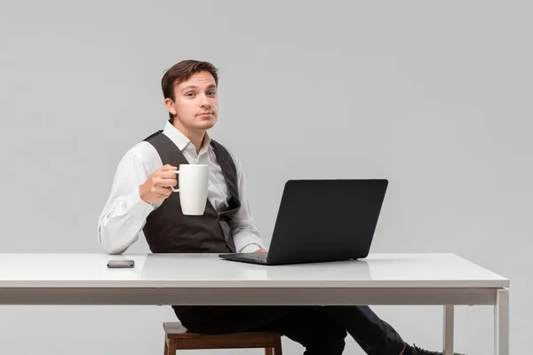 Успішний чоловік у білій футболці та сірому жилеті п'є каву, сидячи за білим столом — стокове фото