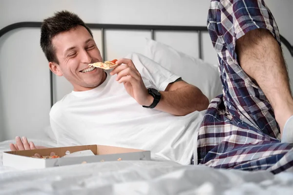 Young man in the white shirt and pajama smiling lying on a bed and eating tasty pizza.