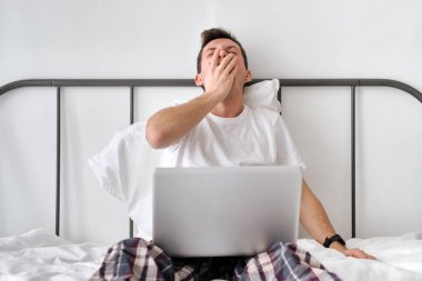 Smiling handsome young man in a white shirt and pajama sitting on a bed with laptop and stretching