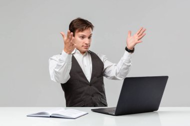 Beyaz tişörtlü ve gri yelekli bir adam beyaz masadaki dizüstü bilgisayara bakarken şok oluyor.