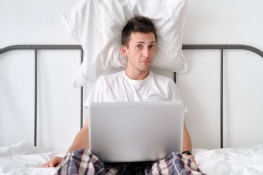 Tired man in a white shirt and pajama sitting on a bed with a pillow behind back and laptop on knees