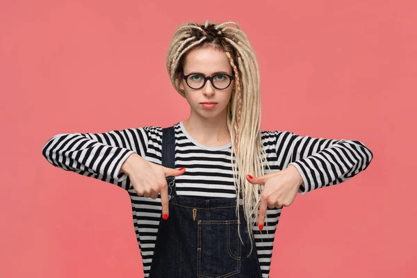 Beautiful young girl with dreads in a striped shirt and jeans jumpsuit pointing at copy space down — ストック写真