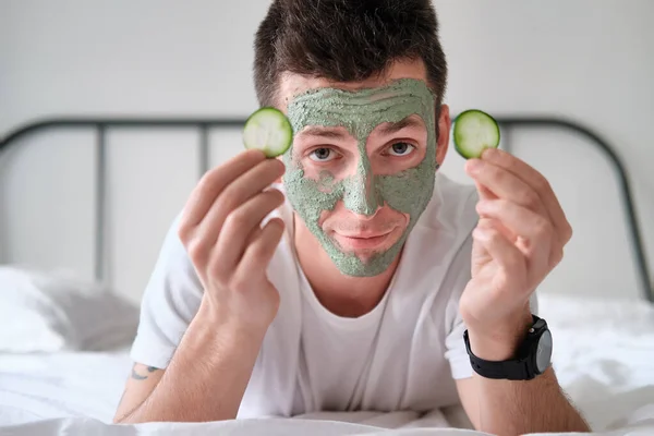 Young man in a white shirt with applied green cosmetic mask holding pieces of cucumber