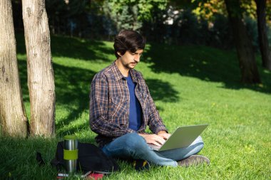 Bıyıklı, sakallı, esmer bir öğrenci çimlerin üzerinde dizüstü bilgisayarla oturuyor. Uzaklık öğrenimi
