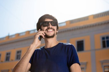Güneş gözlüklü, bıyıklı, sakallı, gülümseyen, cep telefonuyla konuşan genç esmer adam caddede yürüyor. İletişim kavramı