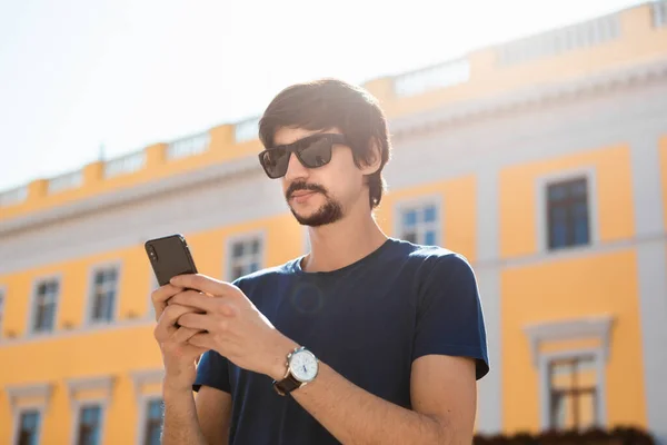 Güneş gözlüklü, sakallı ve bıyıklı genç esmer adam caddede yürüyen bir cep telefonuna bakıyor. İletişim kavramı
