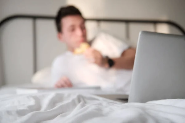 Young man in the white shirt and pajama smiling lying on a bed and eating tasty pizza. Focus on a laptop. Blurred background. Concept of food delivery