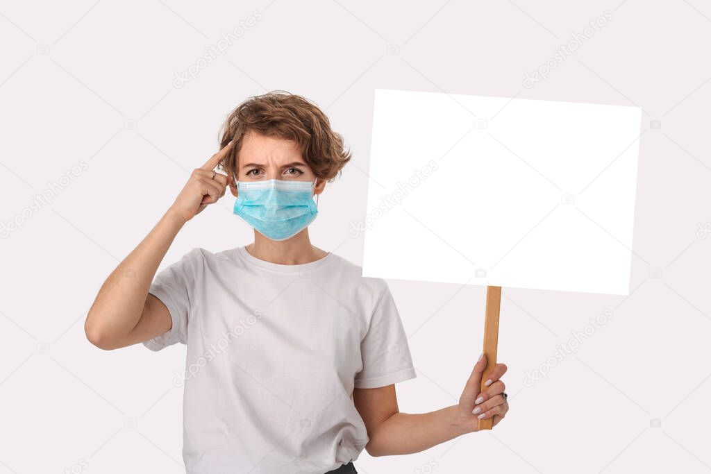 Worried young woman in a white shirt wearing blue medical mask holds picket sign with copy space and piniting at her head. Think about protection from coronavirus.