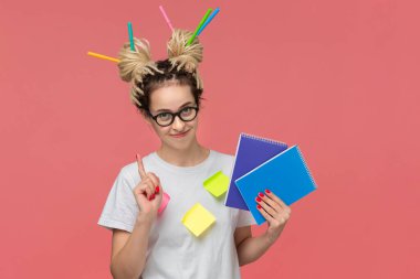 Beyaz gömlekli ve gözlüklü gülümseyen bir öğrenci, bir tişörtte yapışkan notlar ve pembe arka planda izole edilmiş iki defter tutan renkli kalemler. Eğitim ve bilgi kavramı